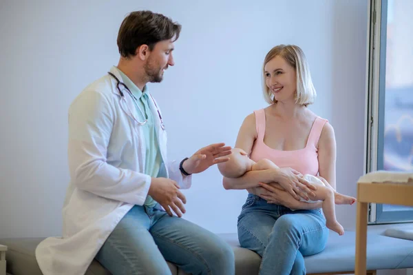 在会诊期间 儿科医师与抱着新生儿的年轻女性家长交谈 — 图库照片