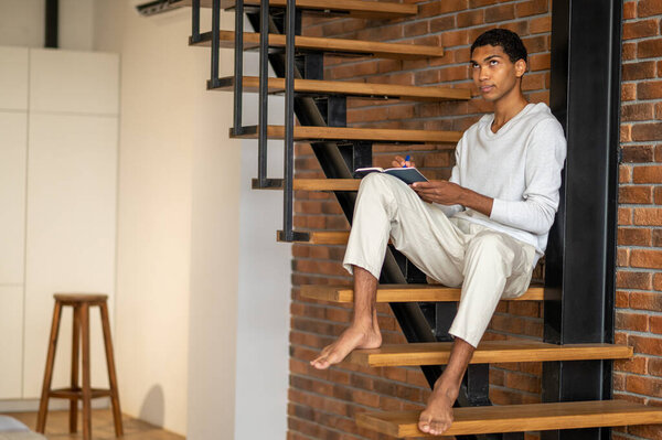 Making notes. Young man in white clothes sitting on wooden stairs and making notes