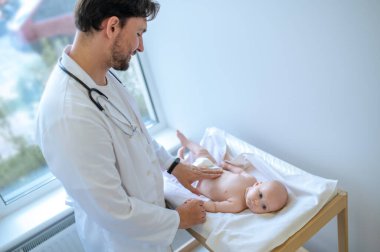 Tıbbi kontrol. Erkek çocuk doktoru bir bebeği muayene ediyor ve olaya dahil olmak istiyor.