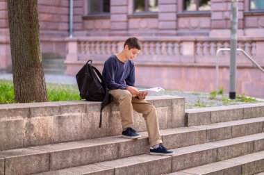 Gülümseyen genç, elinde açık bir kitapla beton merdivenlerde oturuyor.