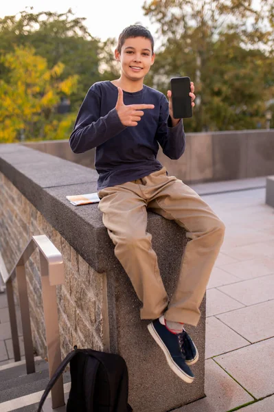Улыбающийся Мальчик Сидящий Бетонных Перилах Указывающий Указательным Пальцем Сотовый Телефон — стоковое фото