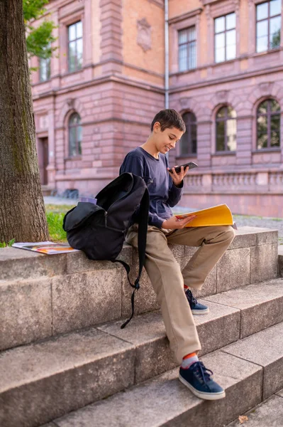 Uśmiechnięty Uczeń Siedzący Betonowych Schodach Otwartą Książką Patrząc Ekran Smartfona — Zdjęcie stockowe