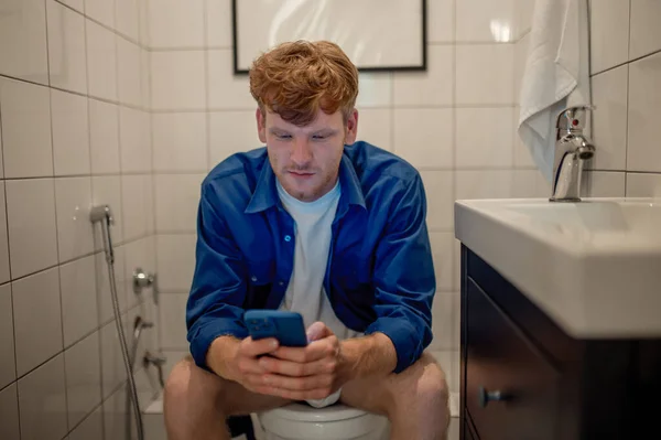 In water closet. Ginger young man sitting in the water closet with a phone in hands