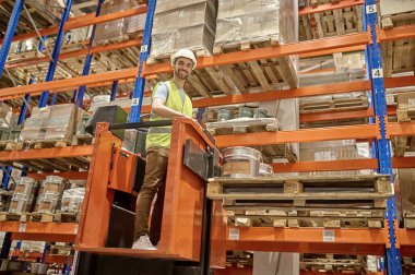 Gülümseyen genç işçi, kaldırılmış forkliftte duruyor. Paslanmaz çelik rafların arasında. Paletlenmiş mallarla.