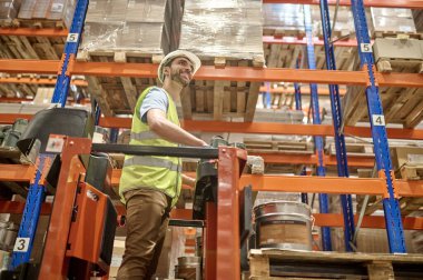 Neşeli bir depo çalışanı kaldırılmış forkliftte duruyor. Paslanmaz çelik rafların arasında. Paletli mallarla birlikte.