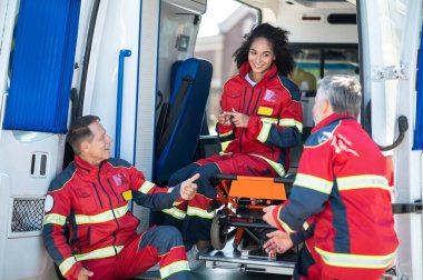 Gülümseyen genç bayan sağlık görevlisi ambulans sedyesinde oturuyor ve iş arkadaşlarıyla konuşuyor.