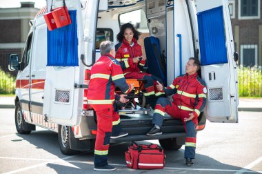 Neşeli Afrikalı Amerikalı kadın sağlık görevlisi ambulans sedyesinde beyaz meslektaşlarıyla sohbet ediyordu.