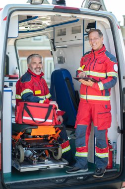 Gülümseyen sağlık görevlisi elinde tablet bilgisayarla ambulans sedyesinde oturan neşeli meslektaşının yanında duruyor.