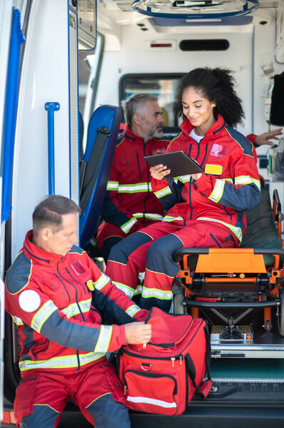 Joyous female paramedic seated on the ambulance gurney using her tablet computer while her concentrated colleagues preparing for work