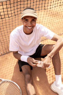 Antrenmandan sonra. Beyaz tişörtlü ve güneşli bir adam tenis kortunda dinleniyor.