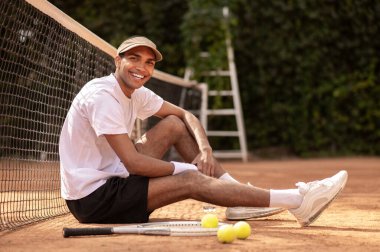 Antrenmandan sonra. Beyaz tişörtlü ve güneşli bir adam tenis kortunda dinleniyor.