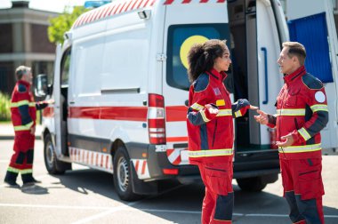 Gülümseyen genç bayan sağlık görevlisi ambulans minibüsünün yanında orta yaşlı erkek arkadaşıyla konuşuyor.