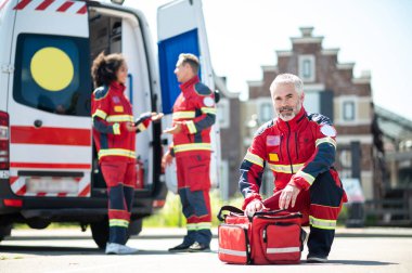 Mature paramedic sitting on his haunches in front of his medical bag while his colleagues chatting near the EMS vehicle clipart