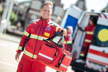 Kırmızı üniformalı neşeli ambulans doktoru bir meslektaşına sağlık çantasını veriyor.