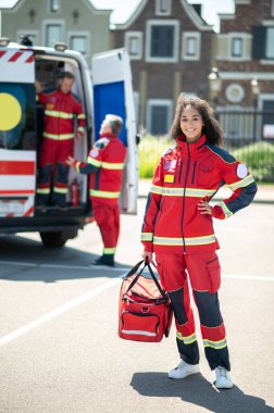 Gülümseyen ambulans doktoru tıbbi çantasını tutuyor ve arka planda meslektaşlarıyla kameraya poz veriyor.