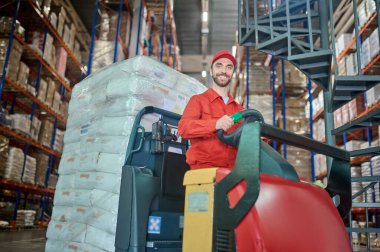 Gülümseyen mutlu depo çalışanı forkliftin direksiyonunda oturuyor ve ileriye bakıyor.