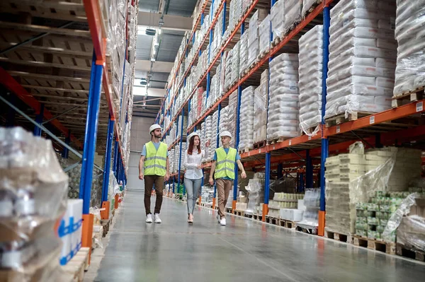 Inspector and storehouse workers walking along a row with the pallet racks loaded with merchandise