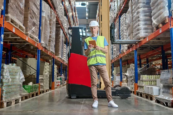 Full-size portrait of a serious warehouse worker with a tablet in the hands standing in the aisle among cargo boxes