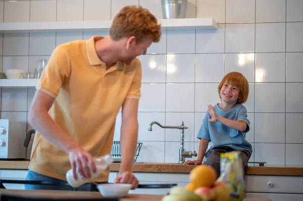 Dad and son in the kitchen. Ginger man with a bottle of milk in hands getting ready the breakfast for his son