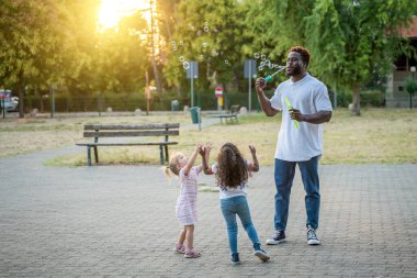 Afrikalı Amerikalı adam iki küçük kızın önünde değnekle yanardöner kabarcıklar üflüyor.