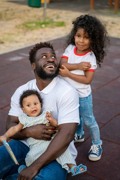 Usmívající Radostný Mladý Afroameričan Sedí Hřišti Obklopen Jeho Roztomilé Dcery — Stock fotografie