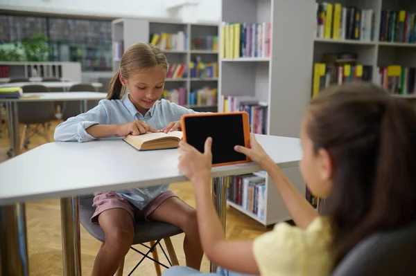 现代学校 女孩们在图书馆里呆着 其中一个用的是智能手机 — 图库照片