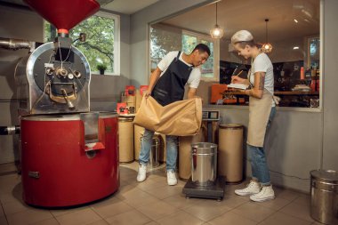 Gülümseyen işçi, neşeli bayan meslektaşı tarafından denetlenen terazinin üzerinde duran kovaya bir şey döküyor.