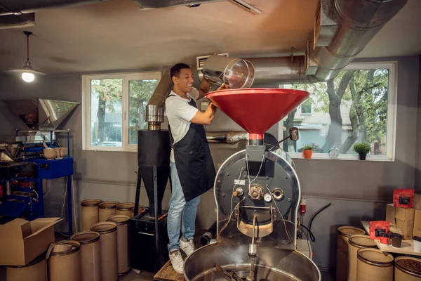 stock image Smiling cheerful young employee pouring the coffee beans from the bucket into the loading hopper