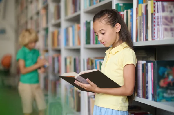 学校图书馆 深色头发的可爱女孩 手里拿着一本书 站在书架上 — 图库照片