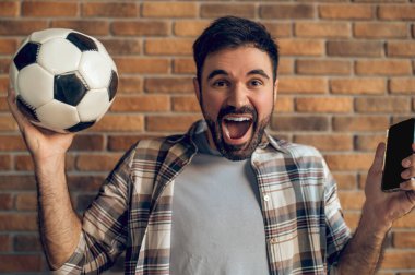 Futbol topu ve cep telefonunu kaldıran ve bağıran duygusal bir adamın portresi.