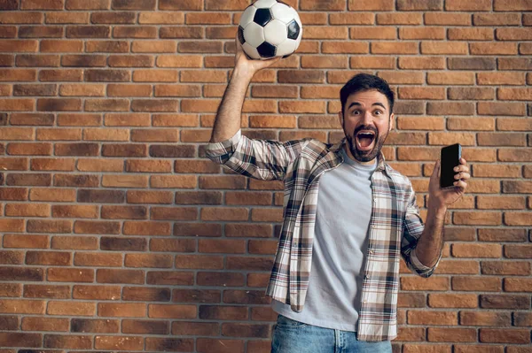 Эмоциональный Любитель Футбола Позирует Камеры Напротив Кирпичной Стены Мячом Смартфоном — стоковое фото