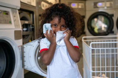 Kıvırcık saçlı küçük kız çamaşır makinesinin yanında tek başına duruyor ve yüzünü kıyafetlerle örtüyor.