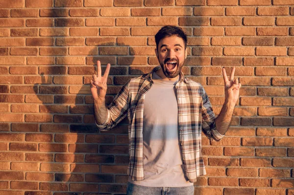 一个快乐的黑头发大胡子男人站在阳光照射下的砖墙旁边 双手做V字形手势 — 图库照片