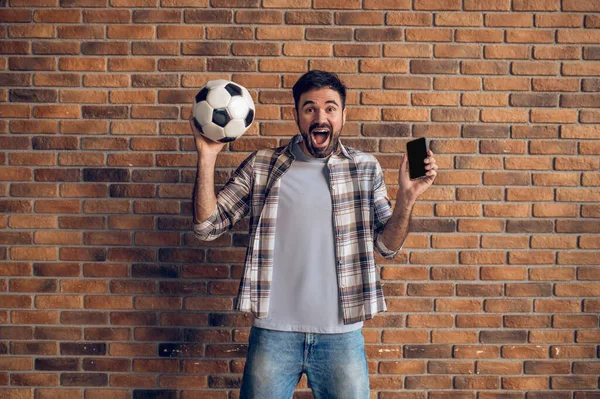 Возбужденный Молодой Человек Держит Футбольный Мяч Смартфон Поднятых Руках Кричит — стоковое фото