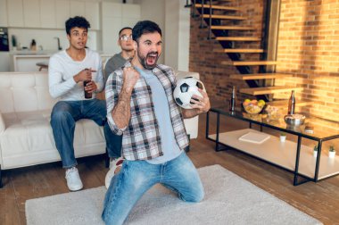 Arkadaşlarının önünde diz çöküp futbol maçı izlerken çığlık atan bir adam.