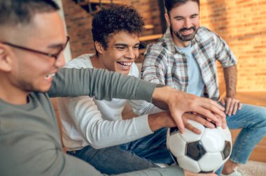 Gülümseyen genç adam ve mutlu arkadaşları ellerini futbol topuna koyuyorlar.