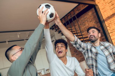 Bağıran neşeli adam ve iki arkadaşı bir futbol topunu havaya kaldırıyorlar.