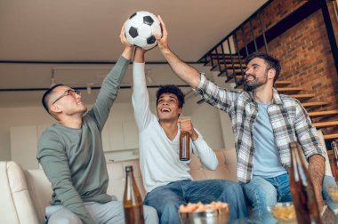 Kanepeye oturmuş futbol topunu havaya kaldıran neşeli adamlar.