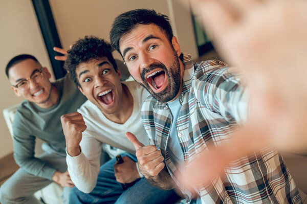 Company of three joyful young men posing for the cellphone camera in the living room