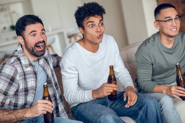 Tres Jóvenes Sorprendidos Sentados Sofá Con Botellas Cerveza Viendo Algo — Foto de Stock
