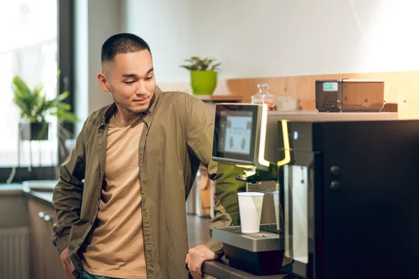 Young Asian corporate worker making himself cappuccino using the coffee machine in the office kitchen