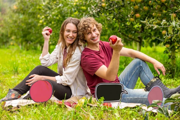 快乐的年轻夫妇 手里拿着苹果 坐在苹果树间的草地上 — 图库照片
