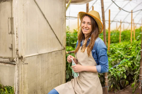 わらの帽子の中で喜びの女性農業者を笑顔とエプロン温室の外にカメラのポーズ — ストック写真