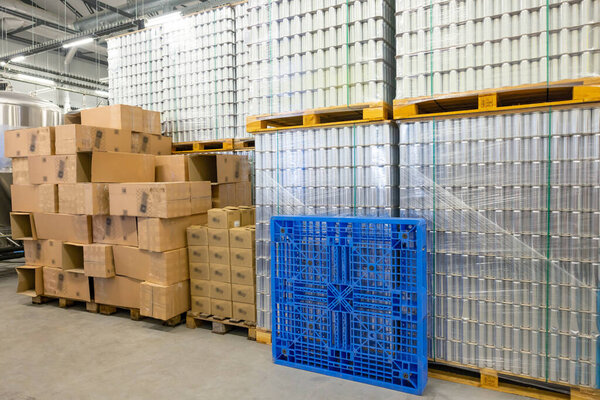 Numerous canned beverages and cardboard boxes stacked on the wooden pallets in a well-lit storage room