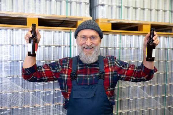 パレットに積み上げられた缶の横に立っている間 彼の手に瓶のペアを保持している陽気な醸造労働者 — ストック写真