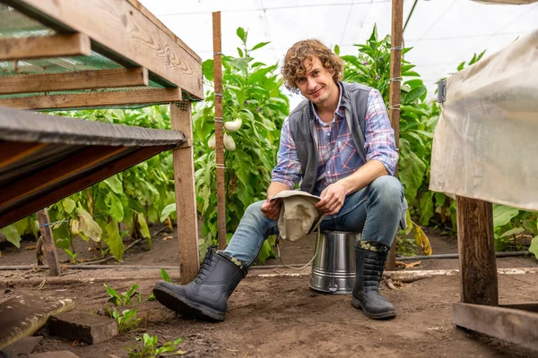 Smiling Agriculturist Holding Hat His Hands Sitting Overturned Bucket Hothouse — Stock Photo, Image
