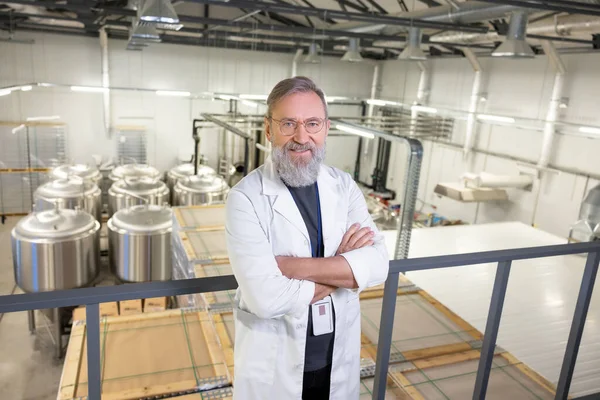 Mature technologist in a lab coat and eyeglasses standing with his arms crossed at a brewery