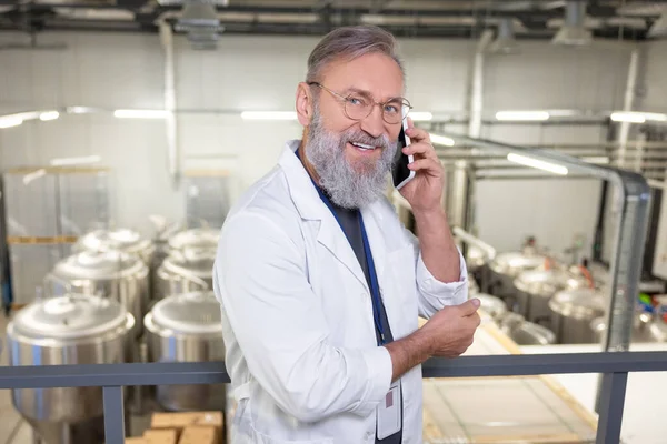 Waist-up portrait of a smiling contented brewery technologist in eyeglasses talking on the smartphone in the factory
