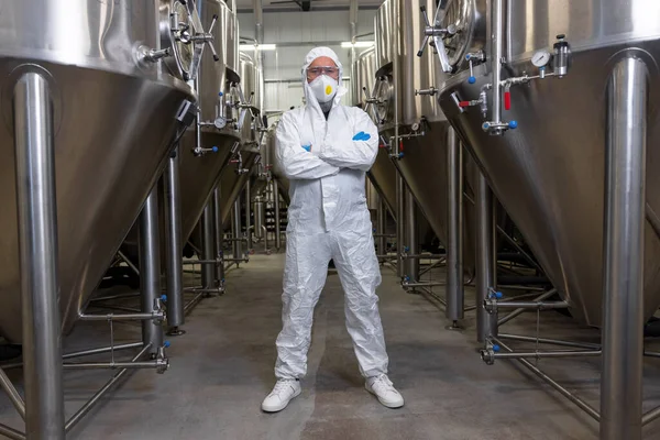 Full-length portrait of a serious brewery technologist in safety clothing standing between the metal tanks in the factory