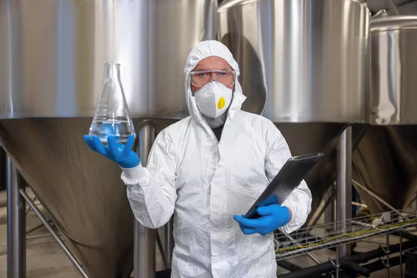 Brewery technologist dressed in protective clothing holding a glass flask with clear liquid in his hands